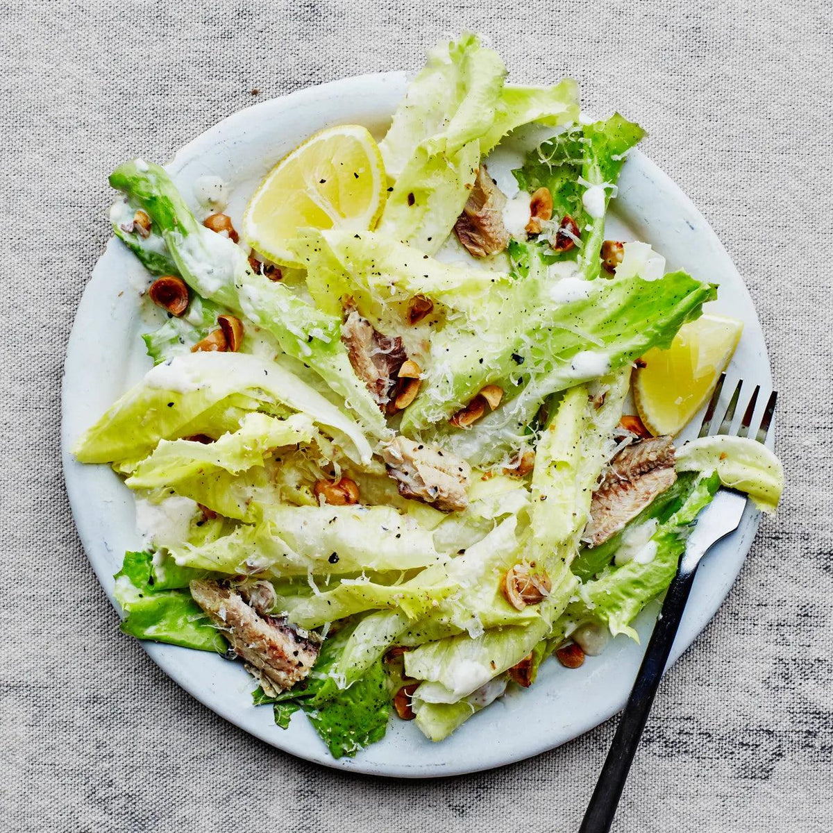 Caesar salad with sardines, crisp lettuce, toasted hazelnuts, grated Parmesan and lemon wedges on a white plate.